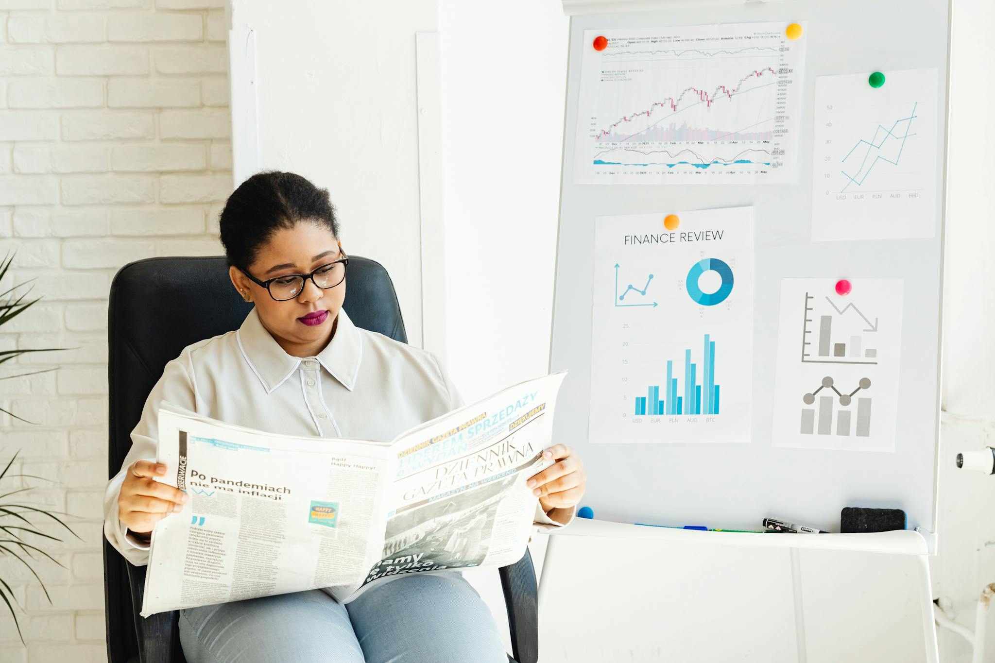 A woman reads a newspaper with stock charts displayed in an office setting.